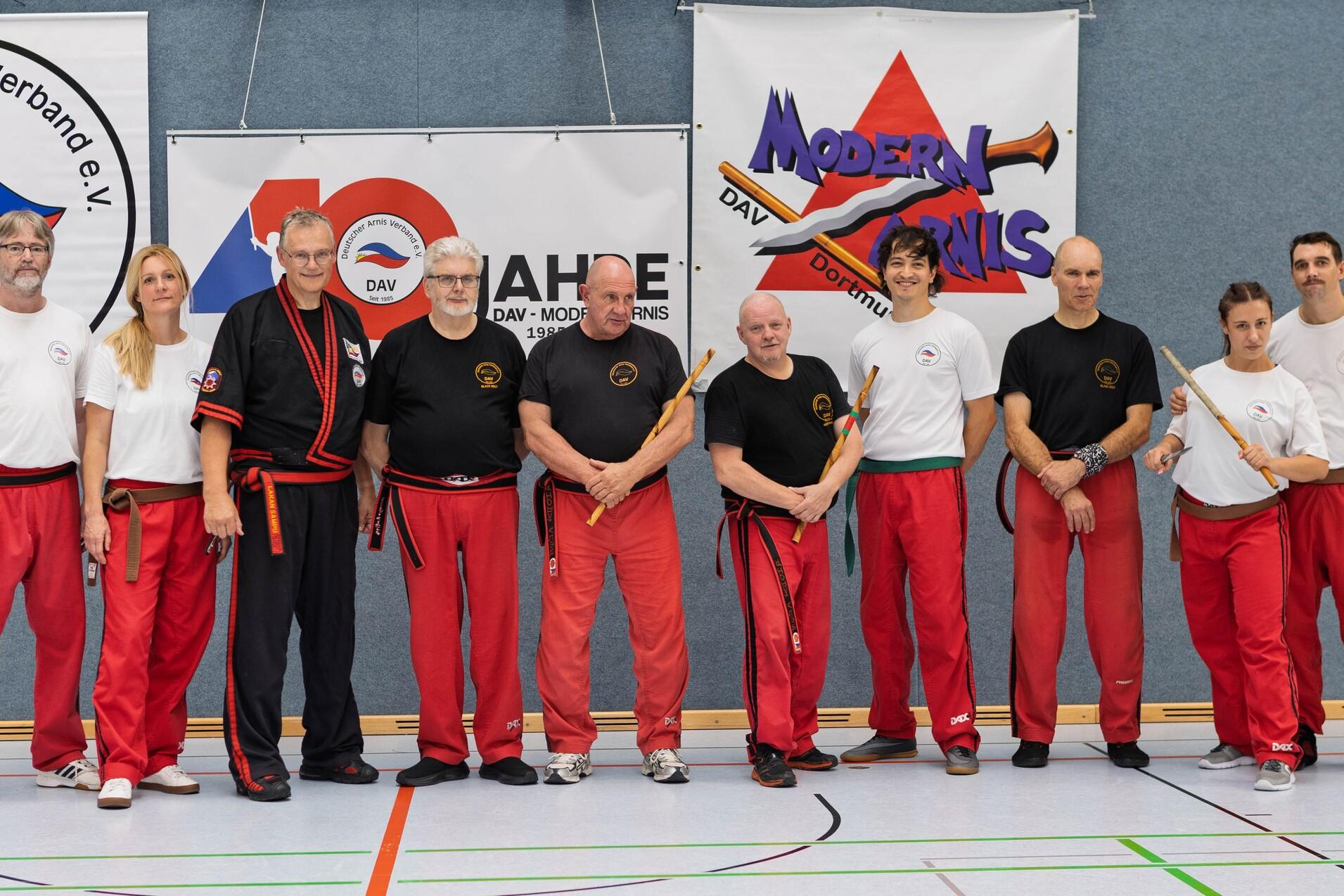 Gruppe von neun Personen in roten Hosen und T-Shirts, posierend in einem Sportstudio vor Bannern des Deutschen Arnis Verbands.
