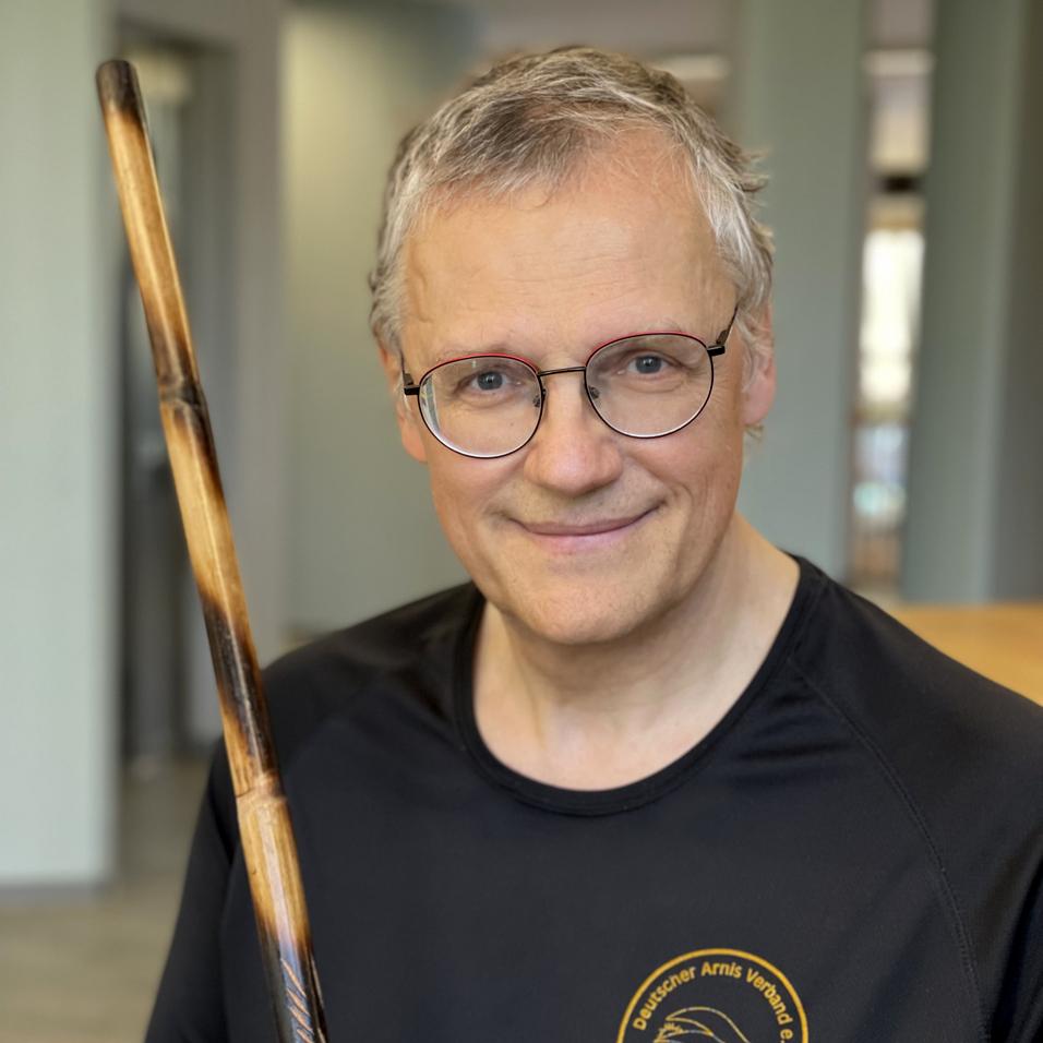 Mann mit Brille hält einen Stock in einem schwarzen T-Shirt des Deutschen Aikido Verbands in einem Innenraum.