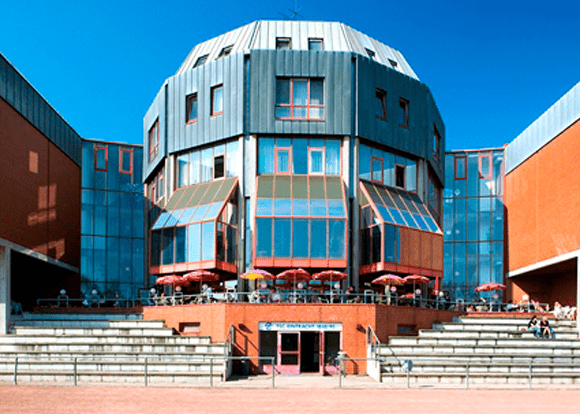 Moderne Architektur eines Gebäudes mit großen Fenstern und Terrasse, umgeben von Treppen und einem blauen Himmel.