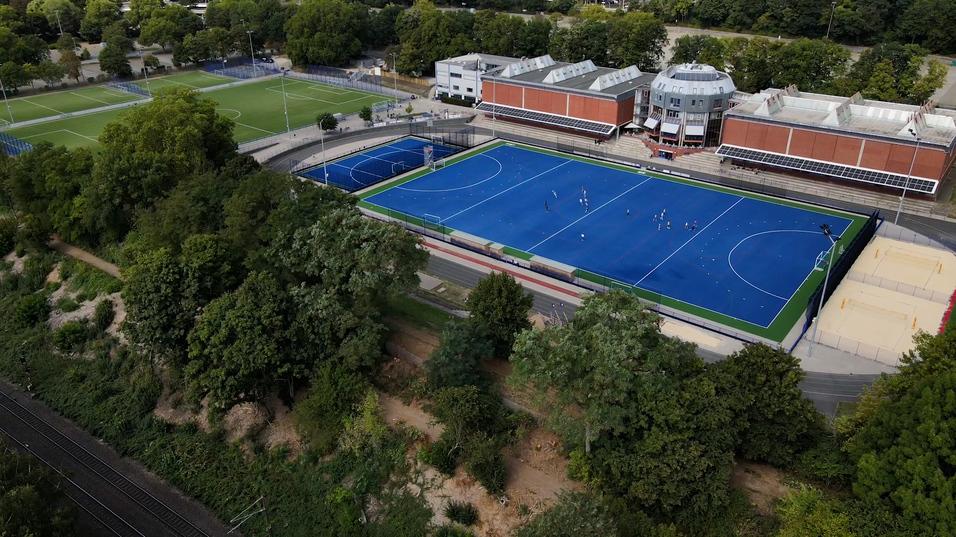 Luftaufnahme eines blauen Hockeyfeldes mit Spielern, umgeben von Bäumen und neben einem Sportgebäude.
