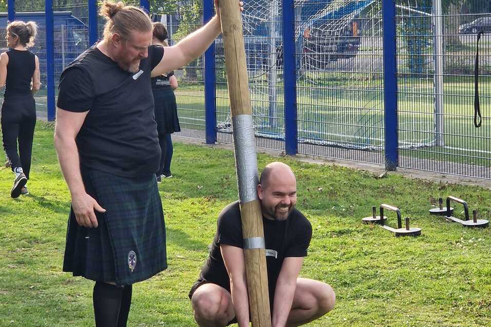 Zwei Männer üben im Freien an einem Holzbalken; einer hält den Balken, der andere beugt sich zum Anheben.