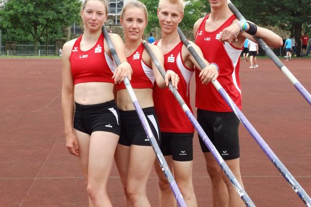 Vier Sportler in roten Trikots stehen auf einer Laufbahn und halten Stabhochsprungstangen, im Hintergrund eine Sportst&auml;tte.