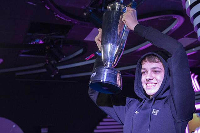 Junger Mann in schwarzem Hoodie hebt einen Pokal in die H&ouml;he und l&auml;chelt vor buntem Hintergrund.