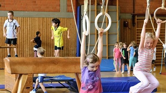 Kinder turnen in einer Turnhalle, einige an Ringen, andere auf einem Schwebebalken und einem Trampolin.