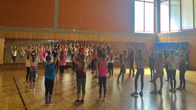 Gruppe von Kindern in einem Sportraum, die mit erhobenen H&auml;nden einer &Uuml;bung folgen.