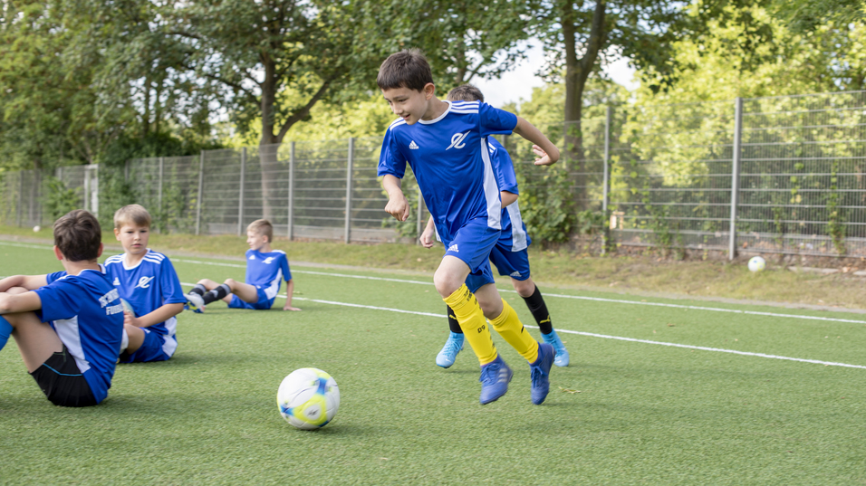 Zwei Jungen in blauen Trikots spielen Fu&szlig;ball auf einem Rasenplatz, w&auml;hrend andere Kinder im Hintergrund sitzen.