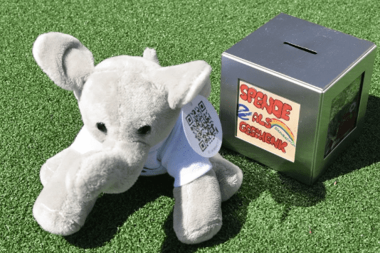 Pl&uuml;sch-Elefant mit QR-Code auf einem gr&uuml;nen Untergrund neben einer silbernen Spendenbox.