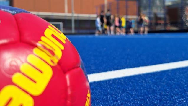 Nahaufnahme eines bunten Handballs auf einem blauen Sportplatz mit unscharfen Spielern im Hintergrund.