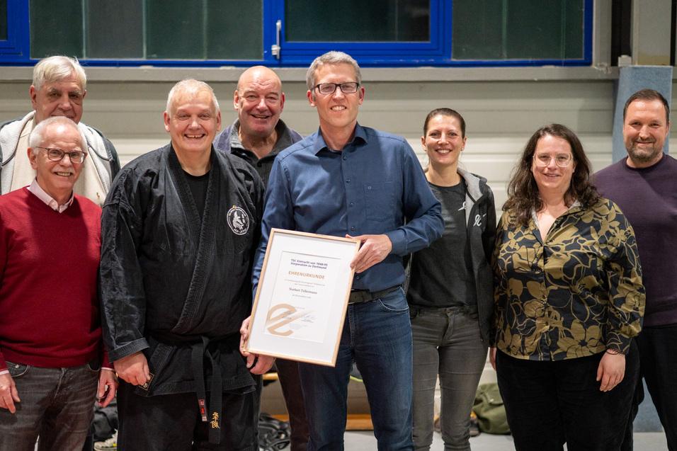 Gruppe von sieben Personen steht l&auml;chelnd zusammen, einer h&auml;lt eine Urkunde in der Hand, in einem Sportraum.