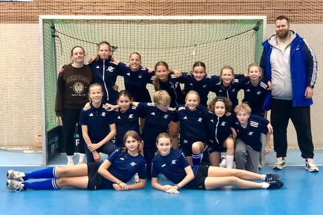 Mannschaftsfoto einer M&auml;dchen-Handballmannschaft mit Trainer vor einem Tor in einer Sporthalle.