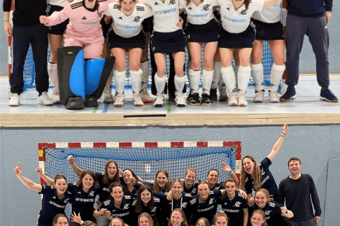 Zwei Gruppen von Frauenhockeyspielern posieren freudig vor Toren in einer Sporthalle.