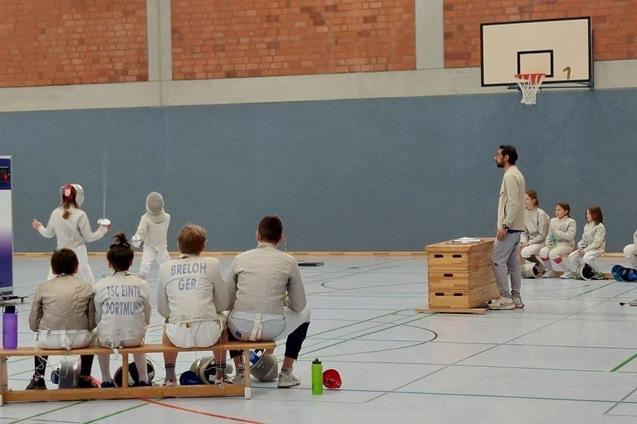 Training einer Fechtgruppe in einer Sporthalle, mit K&auml;mpfern auf der Fl&auml;che und Zuschauern auf einer Bank.