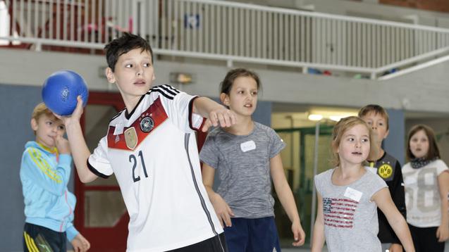 Ein Junge wirft einen Handball, umgeben von mehreren Kindern in einem Sportraum. Alle stehen aufmerksam.