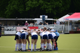 Mannschaftsmitglieder in blau-wei&szlig;en Hockeytrikots bilden einen engen Kreis auf dem Hockeyfeld.