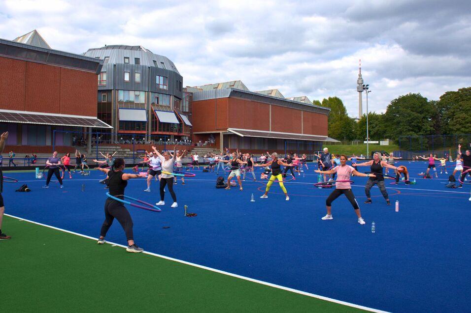 Gruppentraining auf einem blauen Sportplatz, Teilnehmer:innen nutzen Hula-Hoops und Fitnessger&auml;te unter bew&ouml;lktem Himmel.