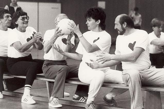 Gruppe von f&uuml;nf Menschen sitzt auf einer Bank, h&auml;lt einen Ball und tr&auml;gt T-Shirts mit Herzmotiven in einer Sporthalle.