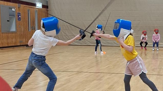 Zwei Kinder im Fechtanzug k&auml;mpfen mit Schwertern in einer Sporthalle, w&auml;hrend Zuschauer im Hintergrund beobachten.