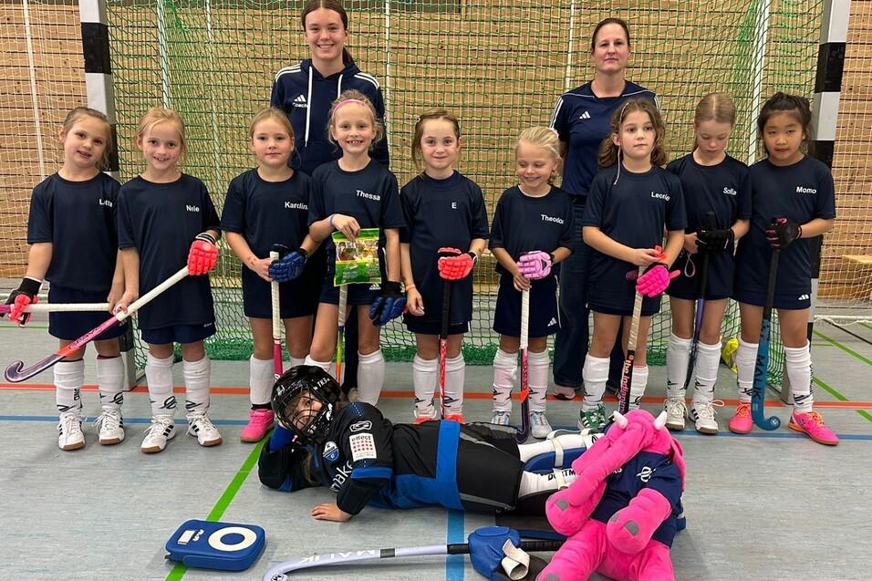 Gruppe von M&auml;dchen in Sportkleidung mit Schl&auml;gern und B&auml;llen, posed vor einem Hockeytor in einer Sporthalle.