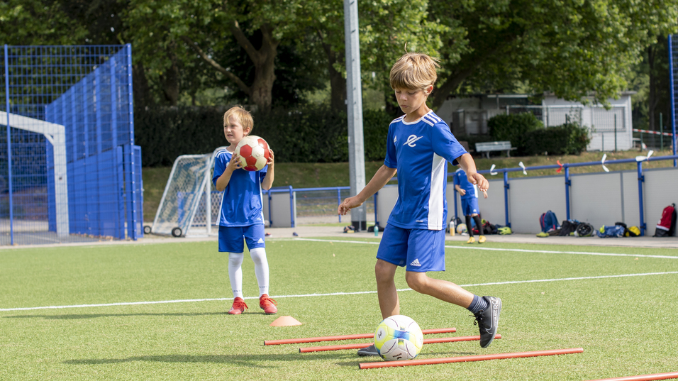 Zwei Kinder in blauen Fu&szlig;balltrikots &uuml;ben auf einem Rasenplatz mit H&uuml;tchen und einem Ball.