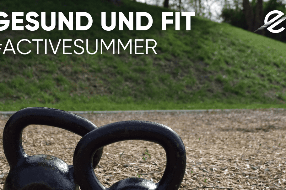 Zwei schwarze Kettlebells liegen auf einem Holzschnitzelboden im Freien. Im Hintergrund ist eine gr&uuml;ne Wiese sichtbar.