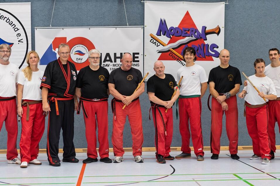 Gruppe von neun Personen in roten Hosen und T-Shirts, posierend in einem Sportstudio vor Bannern des Deutschen Arnis Verbands.