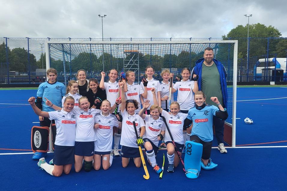 M&auml;dchenhockeyteam in wei&szlig;en Trikots posiert jubelnd vor einem Tor auf einem blauen Kunstrasenplatz.