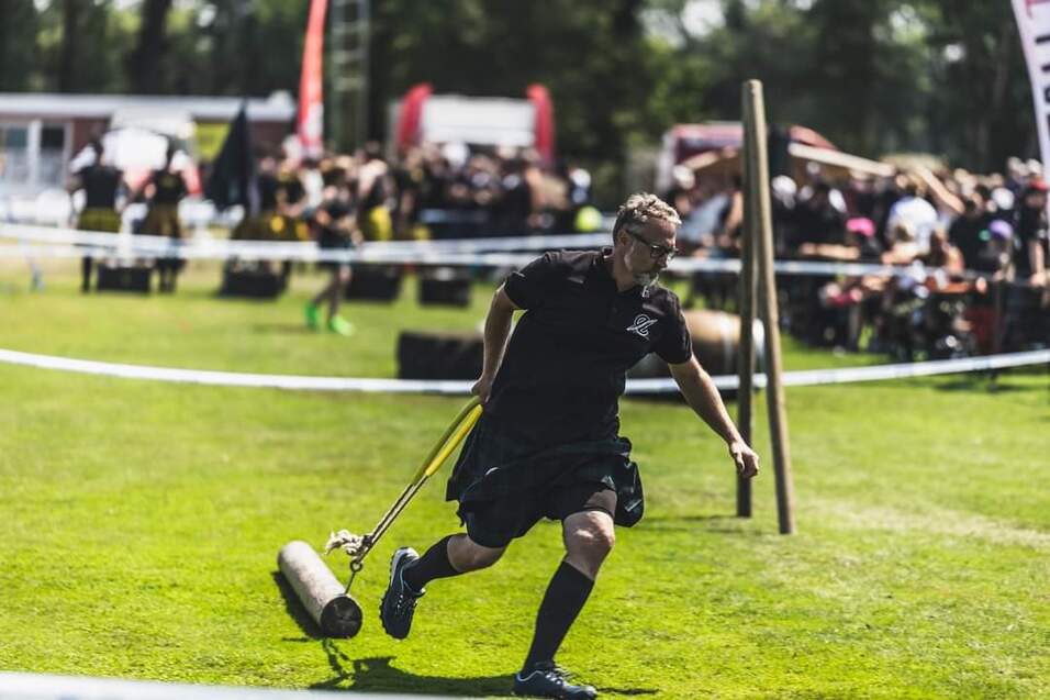 Mann in Schottenrock zieht eine schwere Rolle über ein Grünes Feld bei einem Sportevent. Zuschauer im Hintergrund.