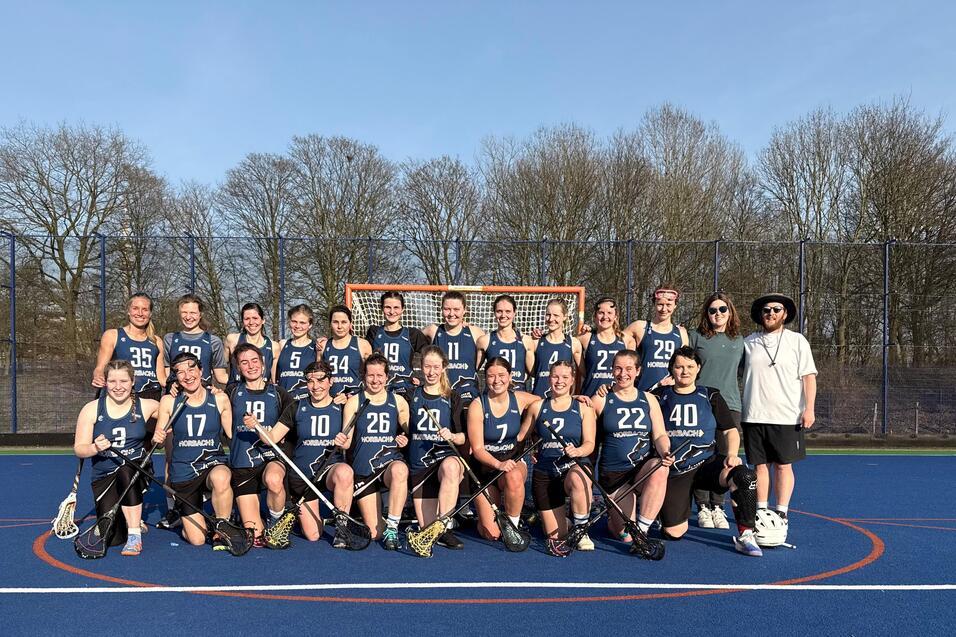 Gruppierung von 22 Lacrosse-Spielerinnen in blauen Trikots auf einem Feld mit Tor im Hintergrund, bei sonnigem Wetter.