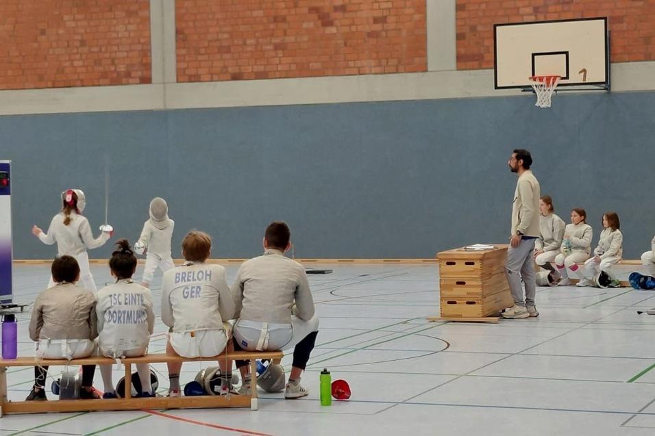 Training einer Fechtgruppe in einer Sporthalle, mit K&auml;mpfern auf der Fl&auml;che und Zuschauern auf einer Bank.