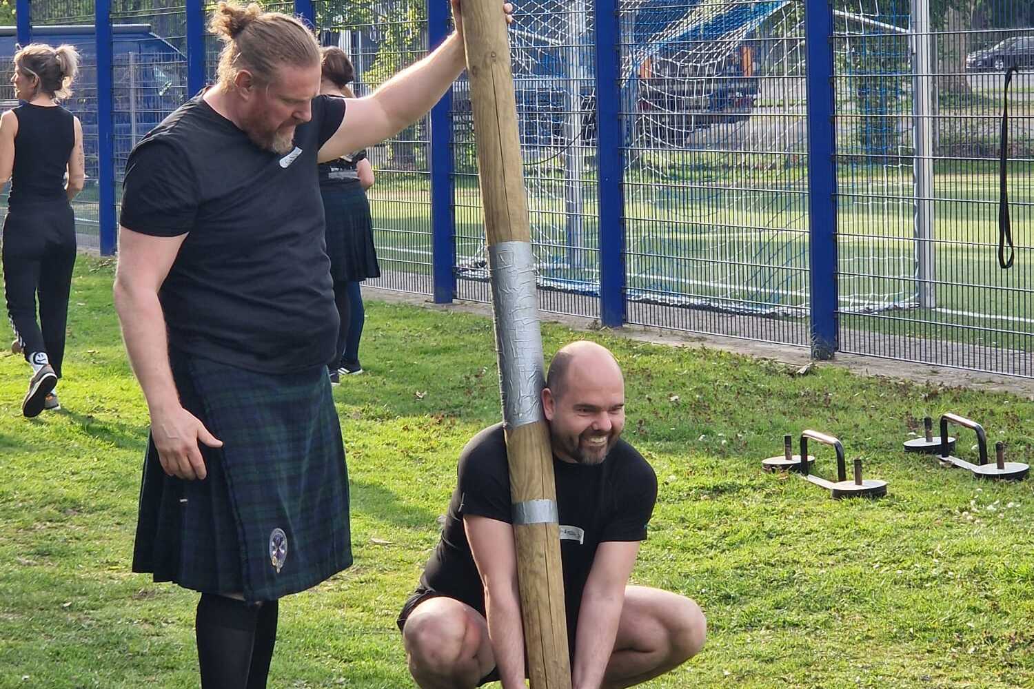 Zwei Männer üben im Freien an einem Holzbalken; einer hält den Balken, der andere beugt sich zum Anheben.