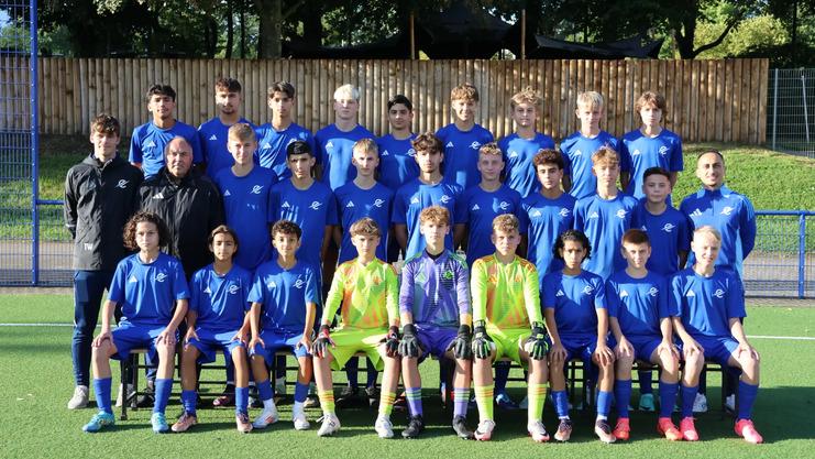 Teamfoto der C1 des TSC Eintracht Dortmund. 28 Personen in drei Reihen auf einem Kunstrasenplatz, alle in blauen Trikots, drei Torhüter in neonfarbenen Outfits. Zwei Trainer außen. Im Hintergrund: Holzzaun und Bäume bei Tageslicht.