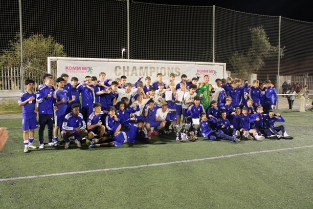 Eine jubelnde Mannschaft blickt direkt in die Kamera. Die Spielerinnen und Spieler lachen, reißen die Arme in die Höhe und zeigen ihre Freude über den Sieg. Vor ihnen stehen mehrere Pokale, die auf ihren Erfolg hinweisen