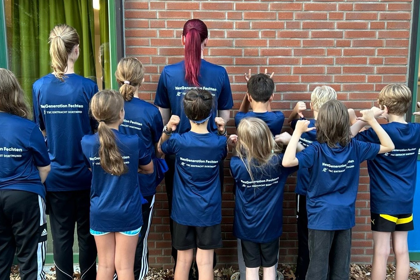 Gruppe von Kindern und einem Erwachsenen in blauen T-Shirts, die von hinten in Richtung einer Wand schauen.