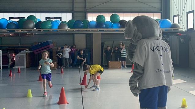 Ein Elefant als Maskottchen beobachtet zwei Kinder beim Hindernislauf in einer Sporthalle, umgeben von bunten H&uuml;tchen.