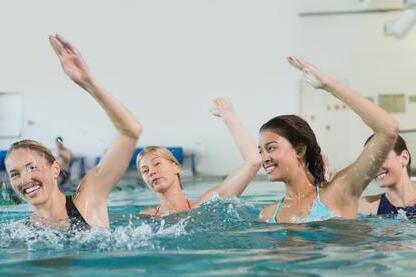 Vier Frauen schwimmen in einem Becken und heben die H&auml;nde fr&ouml;hlich aus dem Wasser.