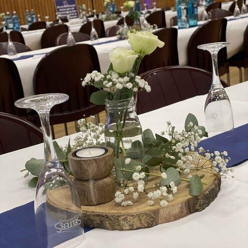 Tischdekoration mit wei&szlig;en Rosen, Eukalyptus und einer Kerze auf einem Holztablett, umgeben von Weingl&auml;sern.