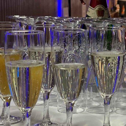 Gl&auml;ser mit sprudelndem Sekt und orangem Getr&auml;nk auf einem Tisch, im Hintergrund unscharfe Personen und Feierdekoration.
