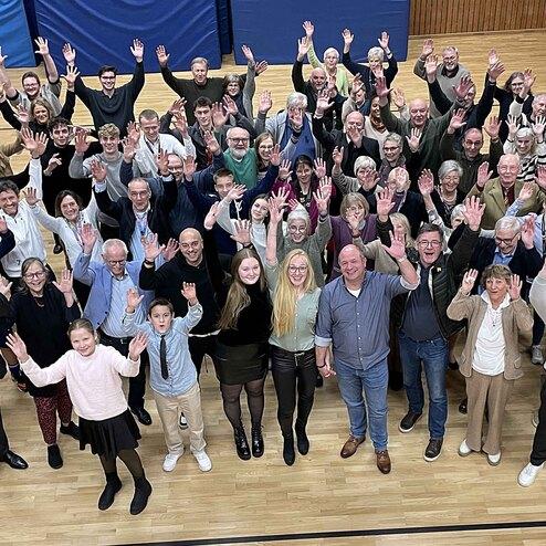 Gruppenfoto von &uuml;ber 100 Personen, die fr&ouml;hlich mit erhobenen H&auml;nden in einem gro&szlig;en Raum stehen.