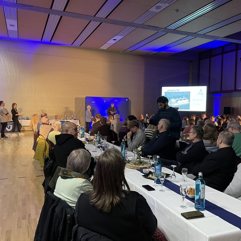 Festliche Veranstaltung mit sitzenden G&auml;sten an langen Tischen, auf einer B&uuml;hne stehen Redner vor einer Pr&auml;sentation.