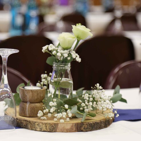 Vase mit wei&szlig;en Rosen und floralen Akzenten auf einem Holzbrett, umgeben von Gl&auml;sern und dekorativen Elementen.