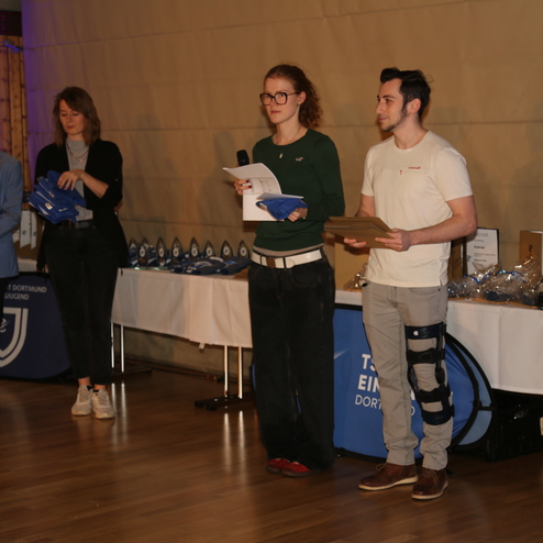 Veranstaltung mit mehreren Personen, die Preise &uuml;berreichen, im Hintergrund eine Tafel mit Ehrungen und Auszeichnungen.