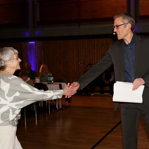 Eine &auml;ltere Frau sch&uuml;ttelt einem Mann die Hand, w&auml;hrend er ein Dokument in der anderen Hand h&auml;lt. Event im Hintergrund.