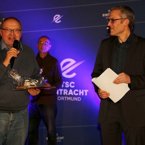 Zwei M&auml;nner stehen auf einer B&uuml;hne; einer h&auml;lt eine Auszeichnung und spricht, der andere h&ouml;rt zu und hat ein Blatt Papier in der Hand.