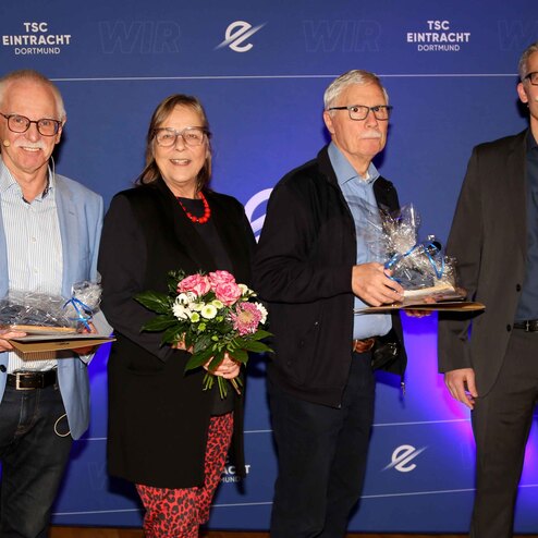 Vier Personen stehen vor einem blauen Hintergrund mit dem Logo des TSC Eintracht Dortmund, halten Urkunden und Blumen.