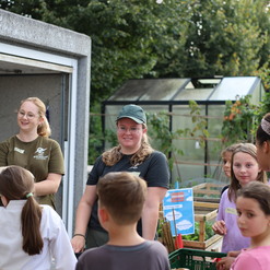 Gruppe von Kindern und Betreuern in einem Garten, einige halten Werkzeuge und schauen interessiert.