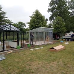 Zwei Gew&auml;chsh&auml;user auf einer Wiese, w&auml;hrend eine Person das transparente Gew&auml;chshaus aufbaut. Werkzeug und Materialien liegen verstreut.
