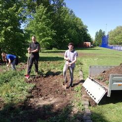 Drei Personen arbeiten gemeinsam im Freien, um einen Garten zu bearbeiten, umgeben von B&auml;umen und einem blauen Zaun.