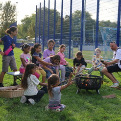 Gruppierung von Kindern und einem Erwachsenen am Lagerfeuer auf einer Wiese, w&auml;hrend sie Stockbrot zubereiten.