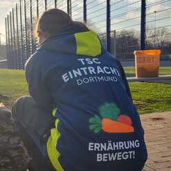 Person in blauer Jacke mit Aufschrift &bdquo;TSC Eintracht Dortmund&ldquo; und &bdquo;Ern&auml;hrung bewegt!&ldquo; sitzt auf einem Platz.