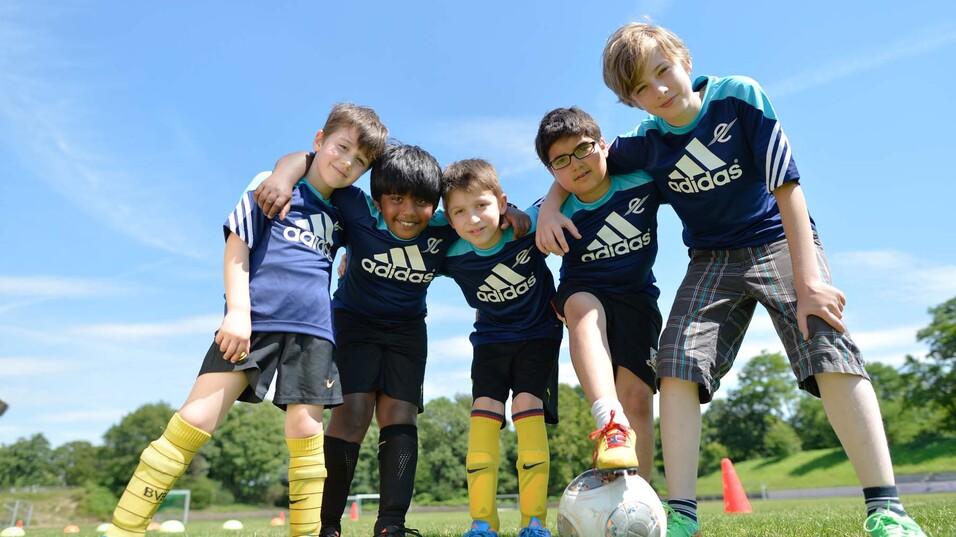 Fünf Kinder in Sportkleidung stehen lachend zusammen mit einem Fußball auf einem grünen Rasenplatz.